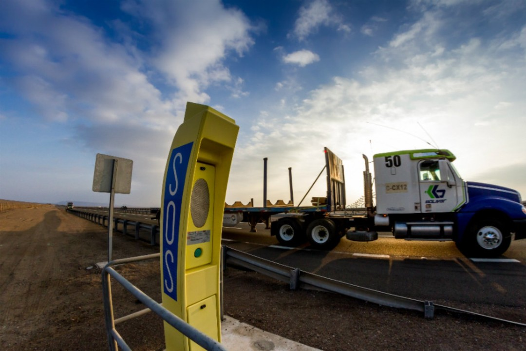 Poste amarillo SOS al costado de una carretera en Antofagasta, con un camión estacionado al fondo y un cielo parcialmente nublado.